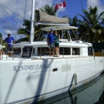 Two men stand on a white catamaran named KINOMOPOLIS docked at a marina.