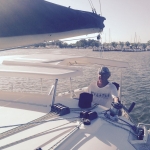 Man steering a sailboat on a sunny day near the shore.