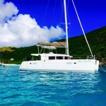 White catamaran floating on clear blue water near green hills under a partly cloudy sky.