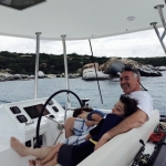 Man and child smiling on a boat steering wheel with ocean in background.