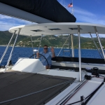 Man steering a sailboat on a sunny day with hills in the background.