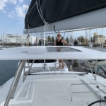 A man stands on a sailboat deck in a marina with boats and masts in the background.
