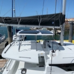 A white catamaran docked at a marina with a covered sail and calm water.